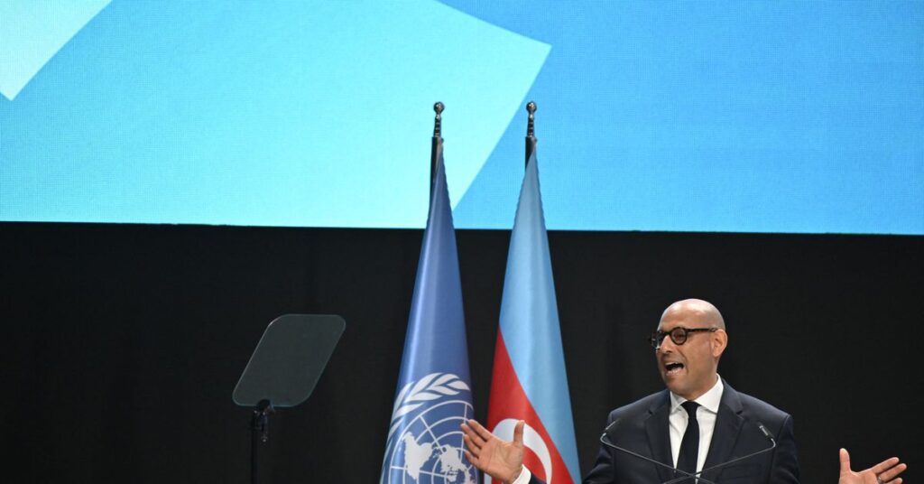 A man wearing a suit and glasses speaks at a podium in front of flags of the United Nations and Azerbaijan.