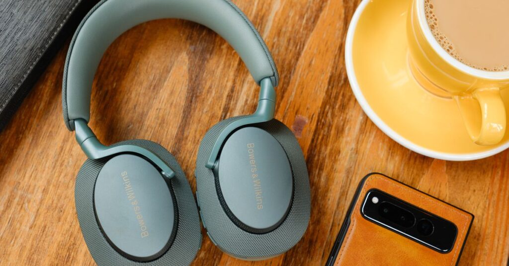 A pair of green Bowers and Wilkins PX7 S2e headphones on a wood table beside a phone and coffee.