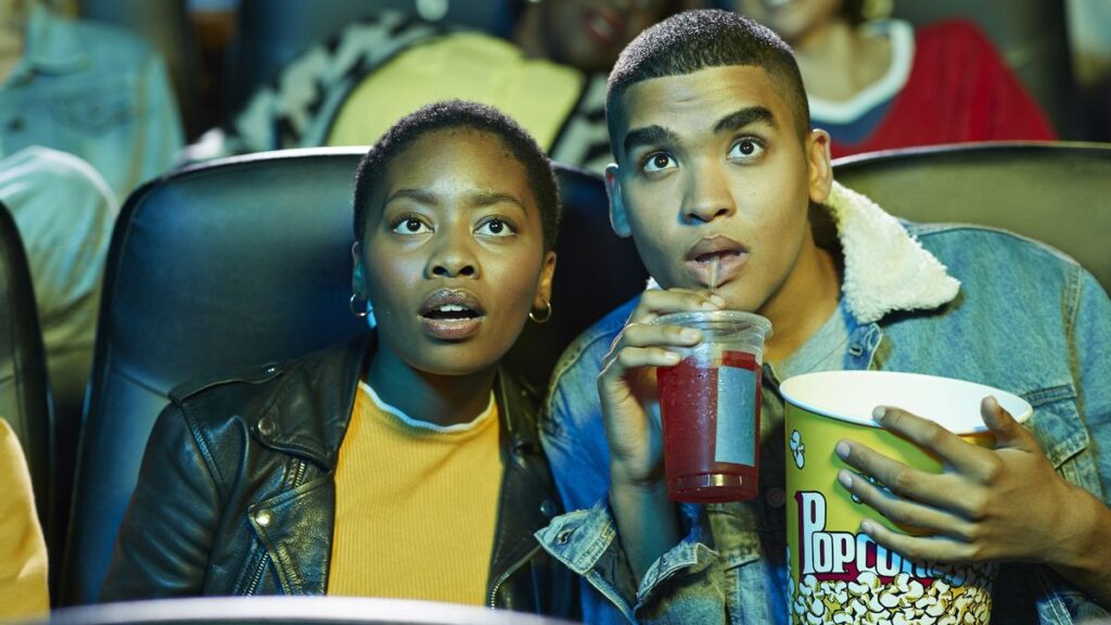 Two young adults lean together as they're watching a movie in a theater; one is holding a popcorn bucket and softdrink