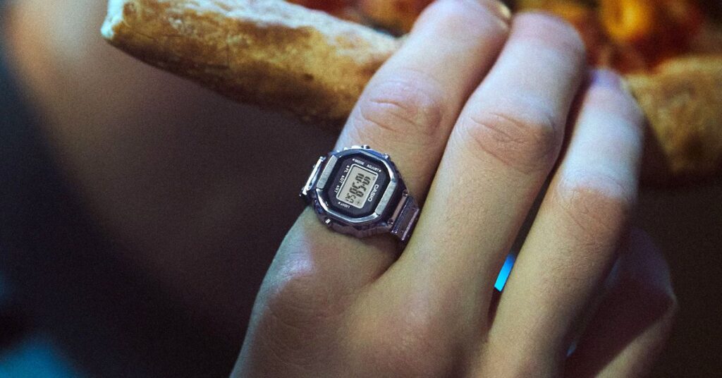 A person holding a slice of pizza while wearing Casio’s functional digital watch ring.