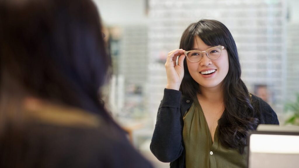 A person with long black hair, a green shirt and black hoodie trying on a pair of clear glasses at a store.