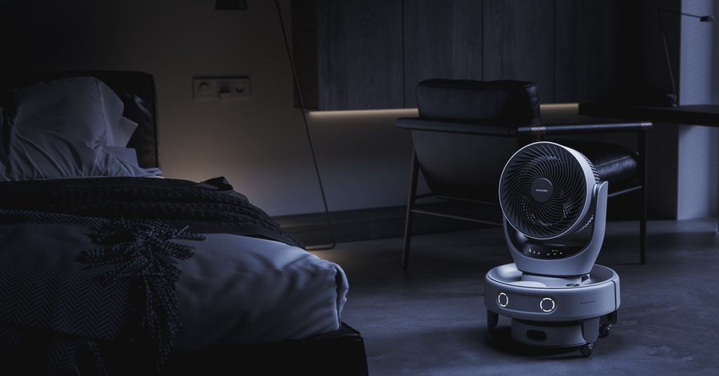 A small white robot with glowing eyes holds a fan next to a bed in a dark bedroom.